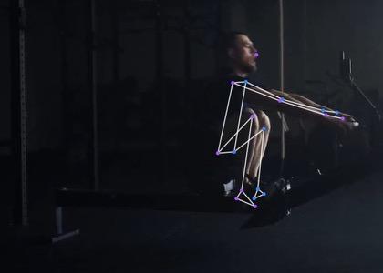 A rower at the catch on an indoor rowing machine, filmed side-on, knees fully bent and arms straight, with the tracked skeleton drawn over the body.